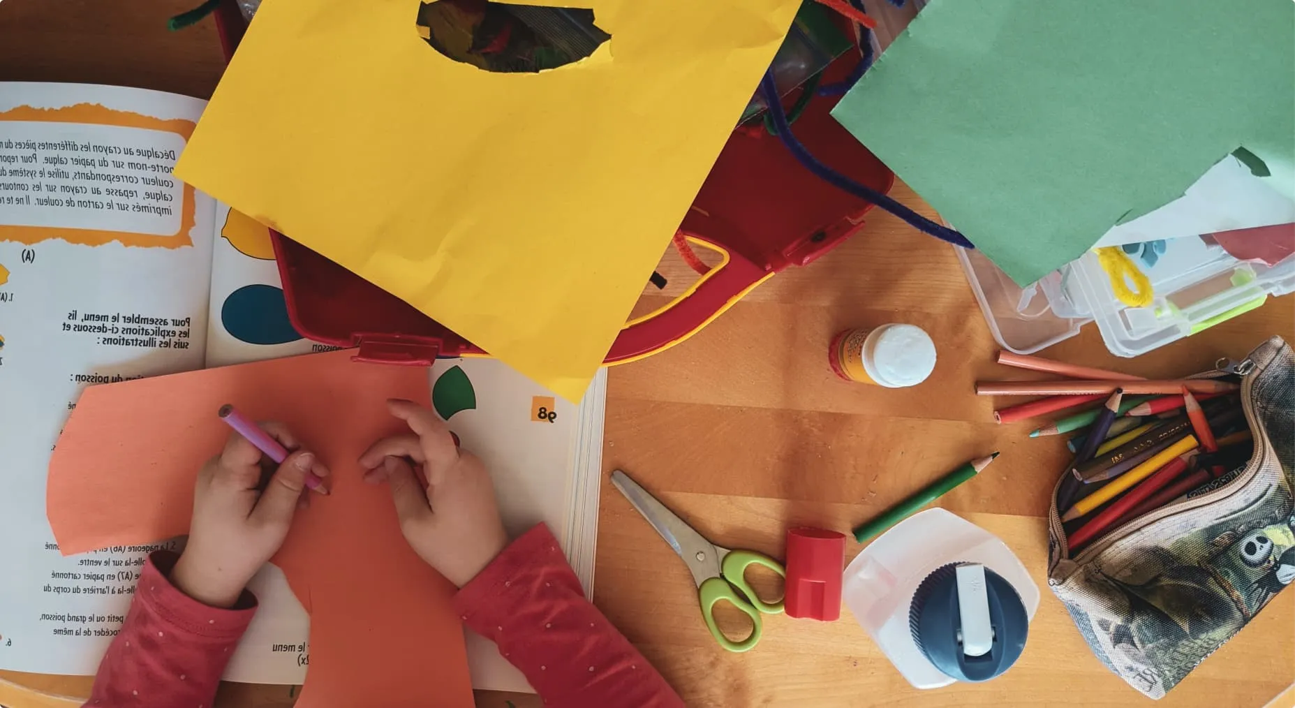 Overhead view of child's hands at a desk full of craft materials for use in online speech therapy, such as scissors, colored pencils, books and more