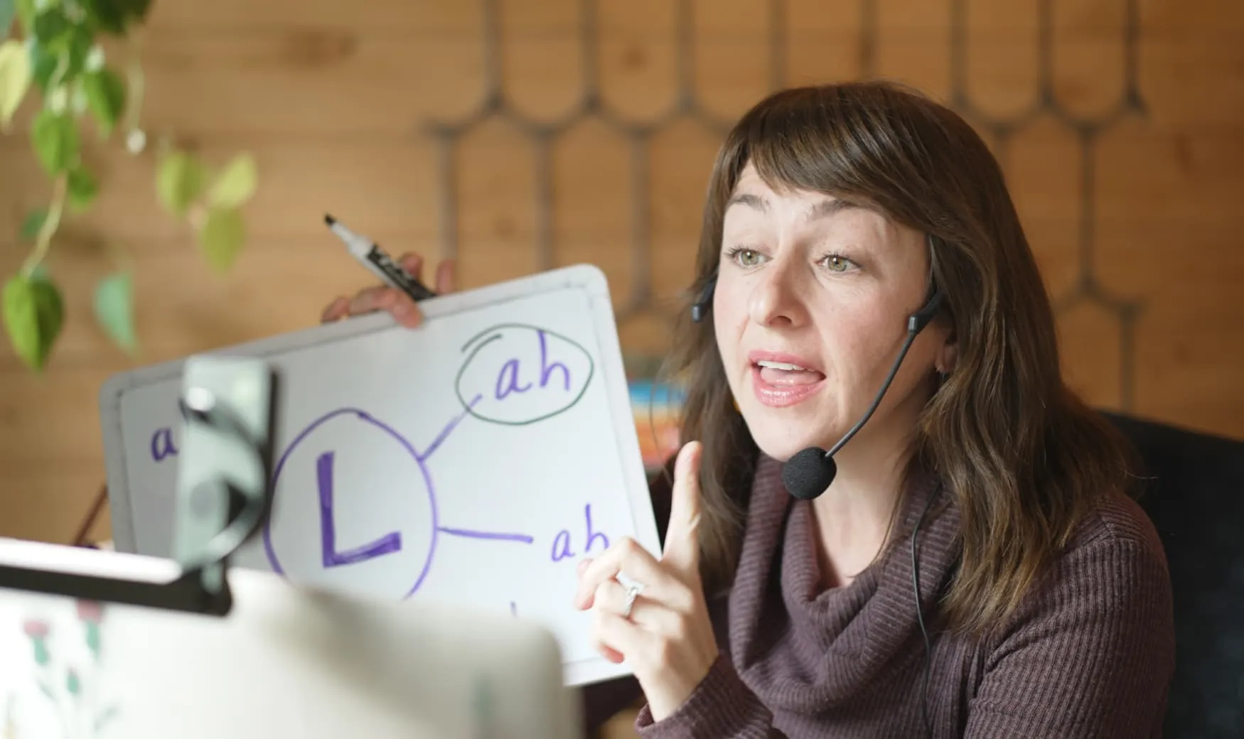 Heather Zortman, wearing a microphone headset, providing online speech therapy. She is pointing to her tongue to model the correct speech sound.